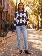 Woman wearing a navy and white argyle crew neck sweater with jeans on a tree-lined street